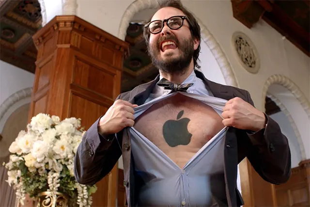 A man in a suit pulling open his shirt to reveal an Apple logo on his chest